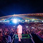 O cantor Cássio Jr se apresenta no palco principal do Festival da Mandioca sob luzes coloridas de show.