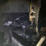 Interior de um cômodo residencial danificado pelo fogo, com paredes manchadas de fuligem e restos de tecidos queimados.