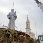 Monumento religioso de grande porte sendo erguido por guindaste no alto de uma colina com vista para a paisagem urbana da capital.