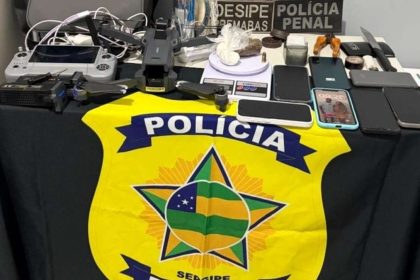Equipamentos eletrônicos, tabletes de maconha, pinos de cocaína e um drone profissional dispostos sobre uma mesa de delegacia como material apreendido.