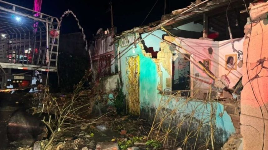 Traseira de um caminhão de carga de botijões de gás cravada na parede destruída de uma casa, com equipes dos Bombeiros e uma retroescavadeira ao redor.