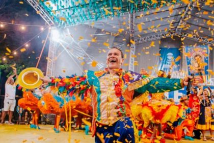 Brincantes da quadrilha junina Século XX sorrindo e dançando com trajes coloridos e caipiras estilizados na arena lotada do Centro de Criatividade.