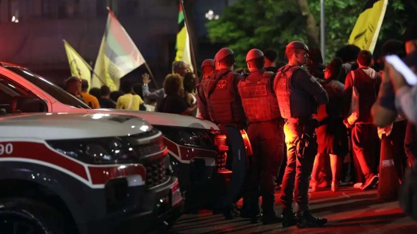 Movimentação de viaturas policiais e peritos em uma comunidade urbana durante a noite, com foco em luzes de emergência.
