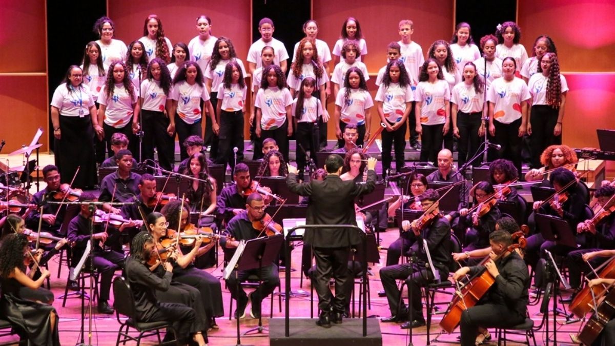 Músicos da Orquestra Jovem de Sergipe tocando instrumentos sinfônicos no palco do teatro.